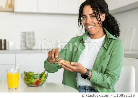 Black lady smiles making sandwich for breakfast sitting at kitchen 110952139