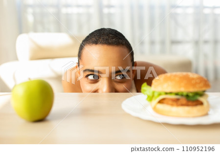 Glad funny millennial latin woman look at hamburger at table with green apple Glad funny millennial latin woman look at hamburger at table with green apple 110952196