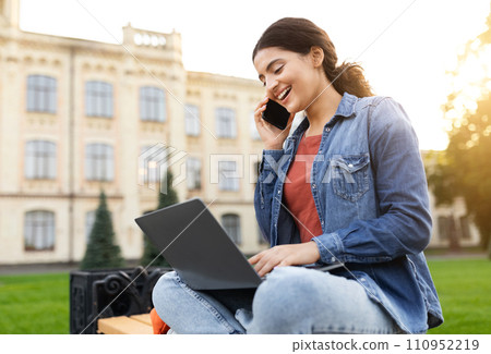 Cheerful young indian lady engaging with laptop, conversing on phone 110952219