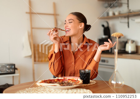 Joyful woman biting pizza slice in a cozy kitchen 110952253