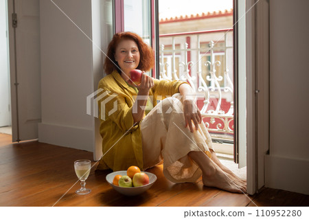 Smiling senior woman sitting by open balcony door and eating ripe apple Smiling senior woman sitting by open balcony door and eating ripe apple 110952280