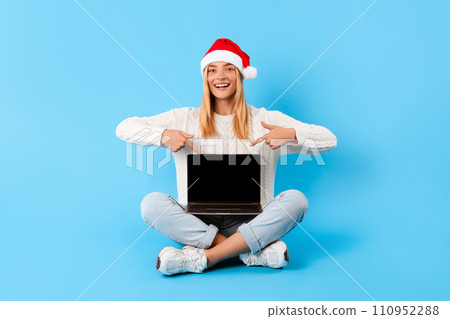 Cheerful lady in Santa hat pointing at laptop screen, blue background 110952288