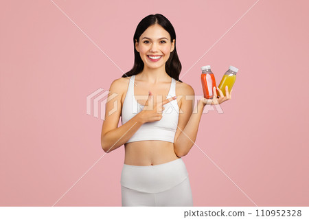 Positive caucasian lady in sportswear holding and pointing at jars of juice or smoothies isolated on pink background Positive caucasian lady in sportswear holding and pointing at jars of juice or smoothies isolated on pink background 110952328