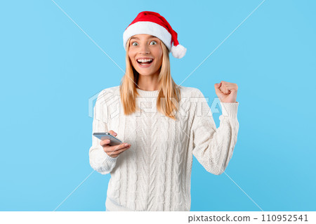 Excited woman in Santa hat seeing Christmas sale on phone and gesturing yes 110952541