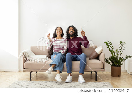 Excited eastern spouses sitting on couch, pointing up Excited eastern spouses sitting on couch, pointing up 110952647