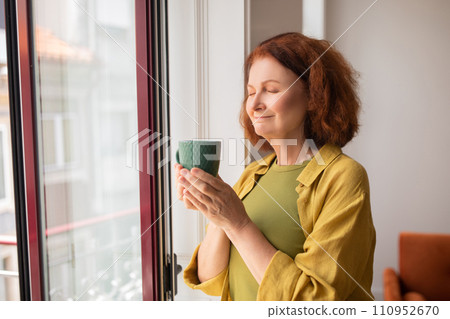 Peaceful senior woman inhaling tea aroma while standing near window at home Peaceful senior woman inhaling tea aroma while standing near window at home 110952670