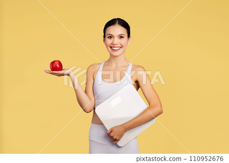 Cheerful caucasian lady in sportswear hold scales, recommend fresh apple isolated on yellow background, studio shot 110952676