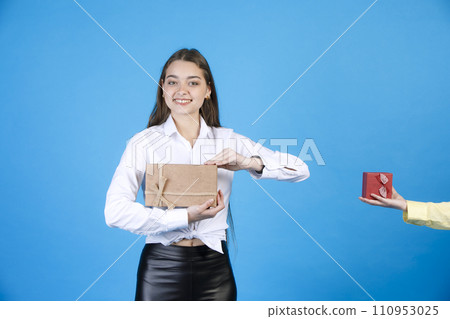 Happy long haired girl enjoying gift, while standing indoors. Portrait of cheerful lady holding present box, unfolding it, while looking at camera, isolated on blue background. Concept of celebration. Happy long haired girl enjoying gift, while standing indoors. Portrait of cheerful lady holding present box, unfolding it, while looking at camera, isolated on blue background. Concept of celebration. 110953025