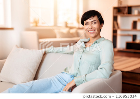 Cheerful middle aged lady sitting elegantly on living room sofa 110953034