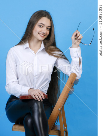 Beautiful happy woman with loose hair posing, while sitting on wooden chair. Portrait of attractive lady holding book on knee, while smiling at camera, isolated on blue background. Concept of posture. Beautiful happy woman with loose hair posing, while sitting on wooden chair. Portrait of attractive lady holding book on knee, while smiling at camera, isolated on blue background. Concept of posture. 110953055