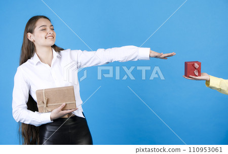 Surprised, pretty birthday girl in elegant shirt, with closed eyes, while holding out arm to receiving wrapped small gift box from unrecognizable male hand, isolated on blue. Concept of expectation. Surprised, pretty birthday girl in elegant shirt, with closed eyes, while holding out arm to receiving wrapped small gift box from unrecognizable male hand, isolated on blue. Concept of expectation. 110953061