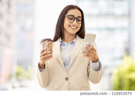 Content businesswoman in a stylish beige jacket holds a coffee cup and smartphone, engaged in reading Content businesswoman in a stylish beige jacket holds a coffee cup and smartphone, engaged in reading 110953074
