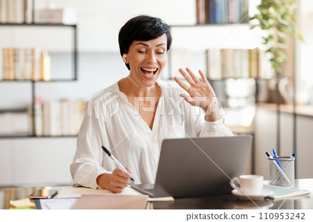 Cheerful middle aged business lady waving hello to laptop indoors 110953242