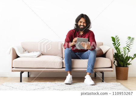 Positive indian man sitting on couch and using digital tablet Positive indian man sitting on couch and using digital tablet 110953267