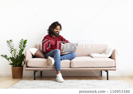 Smiling Indian Man Sitting On The Couch Working On Laptop 110953380