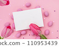 Easter postcard. Invitation mockup or blank with White card for greeting or invitation lies on Easter chocolate pink eggs. Three Red tulips. Shimmering glowing hearts. Happy holidays. Copy space 110953439