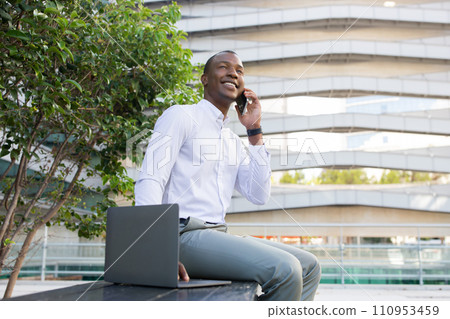 Cheerful black businessman in white shirt having pleasant mobile phone conversation outdoors 110953459