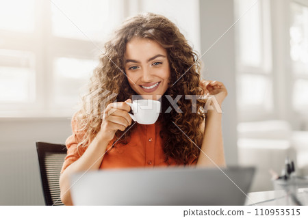 Happy woman with curly hair enjoying coffee at her laptop Happy woman with curly hair enjoying coffee at her laptop 110953515