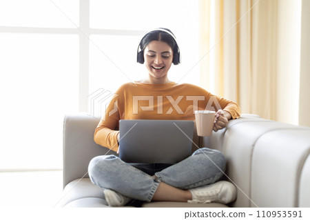 Young Indian Woman Wearing Headphones Resting With Laptop And Coffee On Couch Young Indian Woman Wearing Headphones Resting With Laptop And Coffee On Couch 110953591