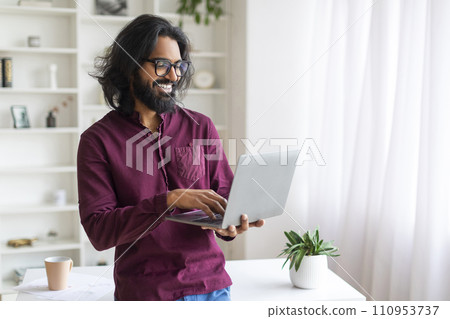 Indian Man Using Laptop While Standing Near Desk At Home, 110953737