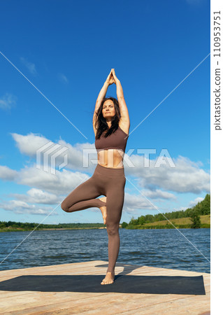 Attractive woman performing Vrikshasana exercise, tree pose, standing with arms raised and joined together, working out in leggings and a short top on the shore of a lake on a warm sunny morning 110953751