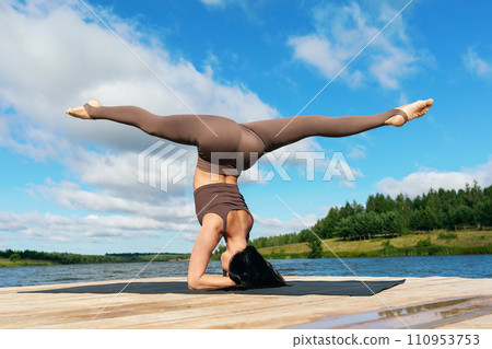 Attractive woman performing Shirsasana exercise with Hanumanasana, inverted asana, headstand with longitudinal splits, training on the shore of a lake on a warm sunny morning 110953753