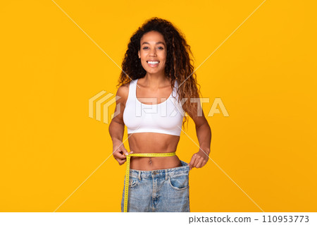 Slim african lady measuring waist with tape over yellow backdrop 110953773