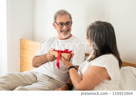 Cheerful senior caucasian woman gives gift box to husband, sit on bad in bedroom interior 110953804