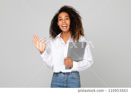 Black millennial lady holding laptop waving hello over gray background 110953883
