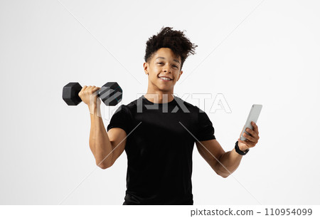 Athletic hispanic man grips dumbbell and smartphone over white background 110954099