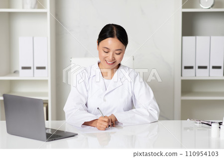 Smiling Asian Woman Doctor Writing Notes While Sitting At Desk In Clinic Smiling Asian Woman Doctor Writing Notes While Sitting At Desk In Clinic 110954103
