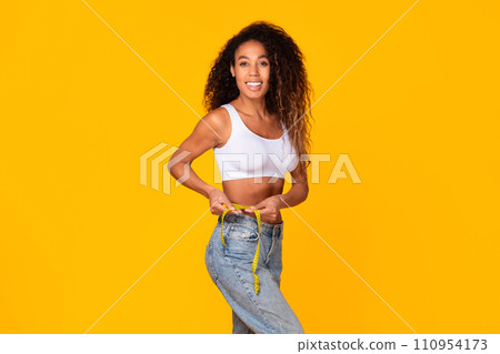 Black lady measuring her waistline with tape, smiling in studio 110954173