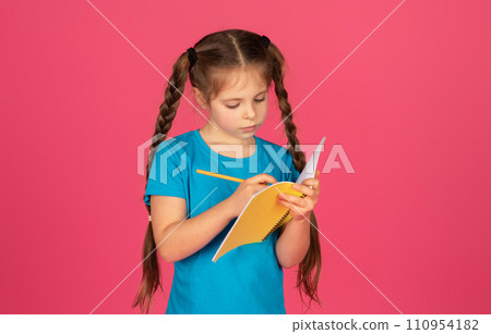 Focused little girl writing in notebook while standing on pink background 110954182