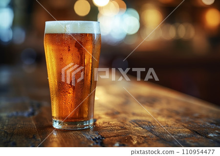 A frosty pint of pale ale, condensation beading on the glass, sitting on a weathered, dark oak vintage table 110954477