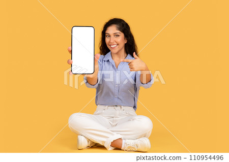 Happy latin young brunette lady point finger at smartphone with empty screen recommend website Happy latin young brunette lady point finger at smartphone with empty screen recommend website 110954496