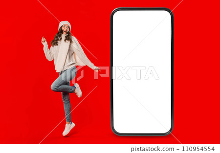 Lady in winter white knitwear poses near huge smartphone, studio 110954554
