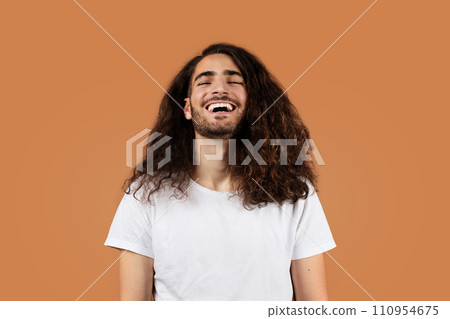 Joyful man with curly brown hair laughs against beige backdrop 110954675