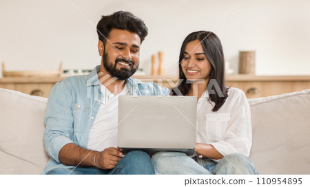 Happy indian couple smiling while looking at a laptop Happy indian couple smiling while looking at a laptop 110954895