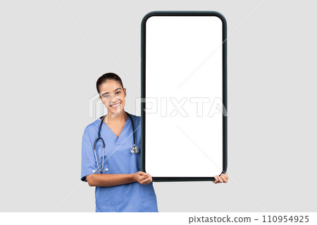 Positive european young lady surgeon or nurse in blue uniform, show big phone with blank screen Positive european young lady surgeon or nurse in blue uniform, show big phone with blank screen 110954925