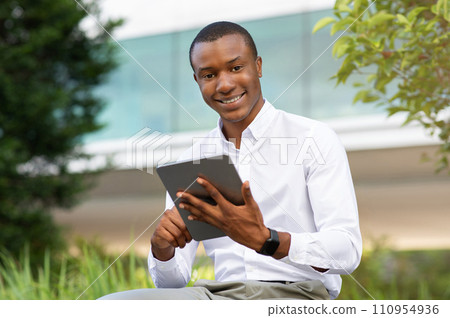 Confident african american businessman using digital tablet while sitting on bench outdoors 110954936