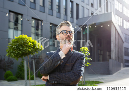 Mature businessman in suit with thoughtful expression standing outside office. Concept of leadership and experience. Mature businessman in suit with thoughtful expression standing outside office. Concept of leadership and experience. 110955525