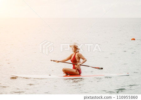 Woman in red bikini on sup board. Happy lady with blond hair in red bathing suit chilling and sunbathing by turquoise sea ocean on sunset. Holiday, vacation and recreational concept. 110955860