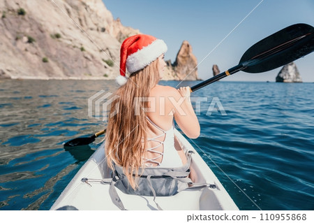 Woman in kayak back view. Happy young woman in Santa hat floating in kayak on calm sea. Summer holiday vacation and cheerful female people relaxing having fun on the boat. 110955868
