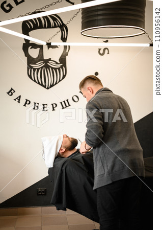 A barber stylist trims the beard of a Caucasian man, whose face is covered with towel, with scissors A barber stylist trims the beard of a Caucasian man, whose face is covered with towel, with scissors 110956142