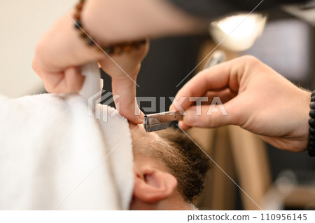 A barber shaves the cheek of a bearded customer with dangerous razor. Shaving the contour of the beard for the correct shape. A barber shaves the cheek of a bearded customer with dangerous razor. Shaving the contour of the beard for the correct shape. 110956145