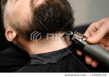A barber in the salon trims the beard of a man with a trimmer in the neck area A barber in the salon trims the beard of a man with a trimmer in the neck area 110956148