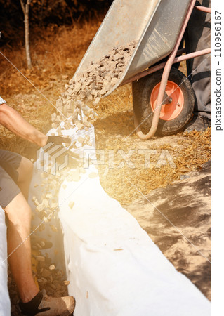 Man pours rubble from a wheelbarrow into a trench. Drainage works for drainage of ground water around the field. Man pours rubble from a wheelbarrow into a trench. Drainage works for drainage of ground water around the field. 110956167