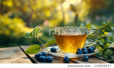 a cup of green tea next to blueberry on a wooden table. blurred background of summer garden from behind a cup of green tea next to blueberry on a wooden table. blurred background of summer garden from behind 110956917