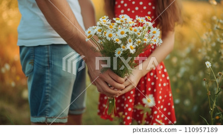 A man in denim shorts and a T-shirt gives a bouquet of daisies to a girl wearing a red polka dot sundress 110957189