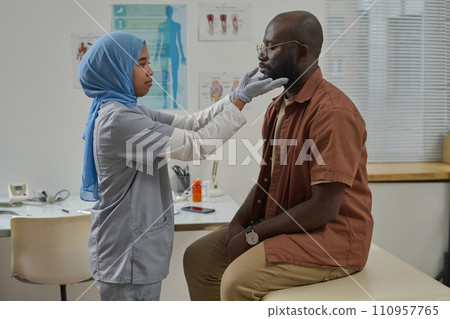 Young muslim doctor in uniform and protective gloves touching lymph nodes of her black male patient Young muslim doctor in uniform and protective gloves touching lymph nodes of her black male patient 110957765